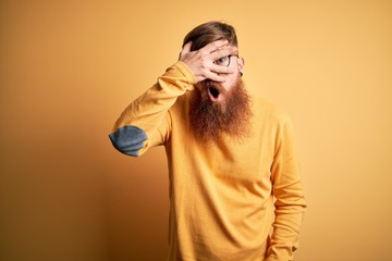 Handsome Irish redhead man with beard wearing glasses over yellow isolated background peeking in shock covering face and eyes with hand, looking through fingers with embarrassed expression.