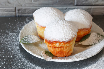Homemade small Easter cakes sprinkled with powdered sugar and decorated with green leaves lie on a white plate on a gray background