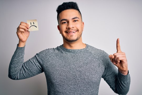 Handsome man holding reminder paper with sad emotion face emoji over white background surprised with an idea or question pointing finger with happy face, number one