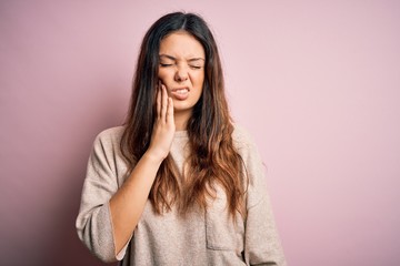 Young beautiful brunette woman wearing casual sweater standing over pink background touching mouth with hand with painful expression because of toothache or dental illness on teeth. Dentist