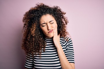 Young beautiful woman with curly hair and piercing wearing casual striped t-shirt touching mouth with hand with painful expression because of toothache or dental illness on teeth. Dentist