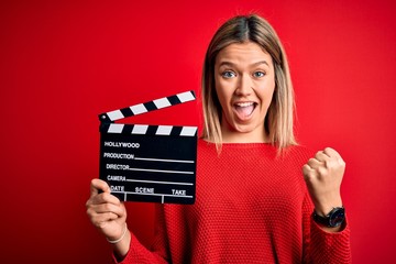 Young beautiful woman holding clapboard standing over isolated red background screaming proud and...