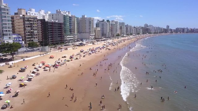Beautiful drone view of "Praia do Morro" beach in Guarapari full of colorful umbrellas in the sand, bathers in the transparent green sea, buildings on the waterfront street and summer blue sky.4K