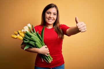 Young blonde woman holding romantic bouquet of tulips flowers over yellow background approving doing positive gesture with hand, thumbs up smiling and happy for success. Winner gesture.