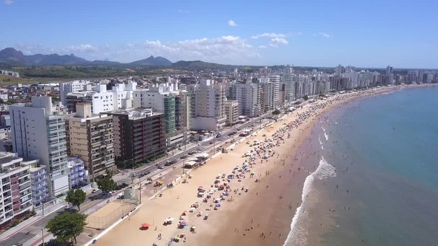 Beautiful drone view of "Praia do Morro" beach in Guarapari full of colorful umbrellas in the sand, bathers in the transparent green sea, buildings on the waterfront street and summer blue sky.4K