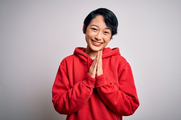 Young beautiful asian girl wearing casual sweatshirt with hoodie over white background praying with hands together asking for forgiveness smiling confident.