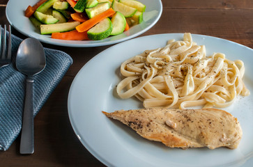 fettuccine and grilled chicken breast with a plate of vegetables on the side on a rustic wooden table