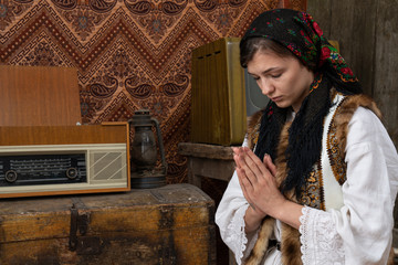 Female in national clothes praying with closed eyes near the aged chest and retro radio