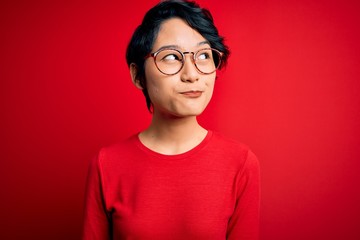 Young beautiful asian girl wearing casual t-shirt and glasses over isolated red background smiling looking to the side and staring away thinking.