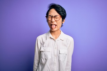 Young beautiful asian girl wearing casual shirt and glasses standing over purple background sticking tongue out happy with funny expression. Emotion concept.