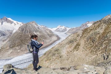 Young Caucasian woman with a backpack on her back, taking a selfie with the monumental Aletsch Glacier behind her.