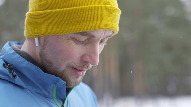 Handheld close up shot of happy bearded man in blue windbreaker and yellow beanie hat wearing wireless earphones and looking down at mobile phone while standing outdoors in winter