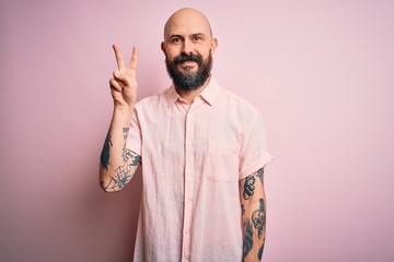 Handsome bald man with beard and tattoo wearing casual shirt over isolated pink background showing and pointing up with fingers number two while smiling confident and happy.