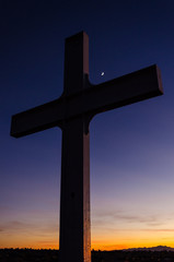 A metal cross under a beautiful, colorful sunset sky and a crescent moon at dusk, Easter