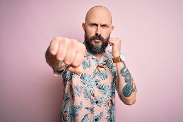 Handsome bald man with beard and tattoo wearing casual floral shirt over pink background Punching...