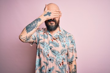 Handsome bald man with beard and tattoo wearing casual floral shirt over pink background covering eyes with hand, looking serious and sad. Sightless, hiding and rejection concept