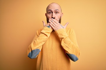 Handsome bald man with beard wearing casual sweater standing over yellow background shocked covering mouth with hands for mistake. Secret concept.