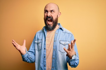 Handsome bald man with beard wearing casual denim jacket and striped t-shirt crazy and mad shouting and yelling with aggressive expression and arms raised. Frustration concept.