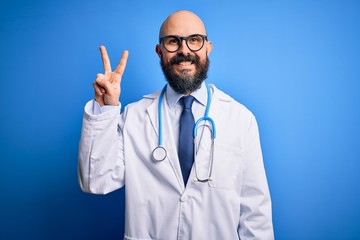 Handsome bald doctor man with beard wearing glasses and stethoscope over blue background smiling looking to the camera showing fingers doing victory sign. Number two.