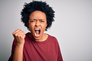 Young beautiful African American afro woman with curly hair wearing casual t-shirt standing angry and mad raising fist frustrated and furious while shouting with anger. Rage and aggressive concept.