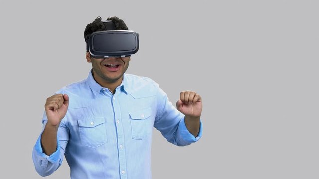Scared hindu man in vr-headset playing horror game. Dark-skinned guy experiencing virtual reality with fear. Grey background.