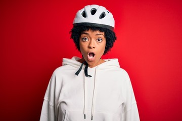 Young African American afro cyciling woman with curly hair wearing bike security helmet afraid and shocked with surprise expression, fear and excited face.