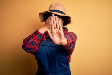 Young African American afro farmer woman with curly hair wearing apron and hat covering eyes with hands and doing stop gesture with sad and fear expression. Embarrassed and negative concept.