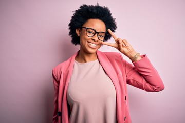 Young beautiful African American afro businesswoman with curly hair wearing pink jacket Doing peace symbol with fingers over face, smiling cheerful showing victory