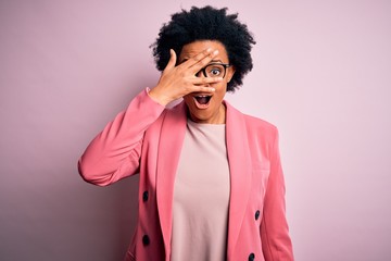 Young beautiful African American afro businesswoman with curly hair wearing pink jacket peeking in shock covering face and eyes with hand, looking through fingers with embarrassed expression.