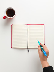 Top view workspace female hand holding blue pen, with free space for writing, cup of coffee, a blank notebook, white background