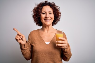 Middle age curly hair woman drinking healthy glass of orange juice over white background very happy pointing with hand and finger to the side