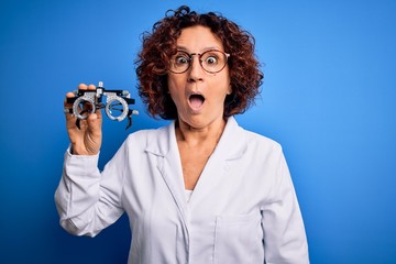 Middle age optical woman wearing coat holding optometry glasses over blue background scared in...
