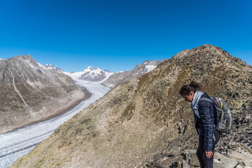 Fototapeta na wymiar Young Caucasian woman with a backpack on her back looking down at the monumental Aletsch Glacier.This giant stream of ice, which stretches over 23 km