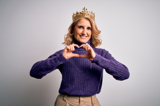 Middle age beautiful blonde woman wearing golden crown of king over white background smiling in love showing heart symbol and shape with hands. Romantic concept.