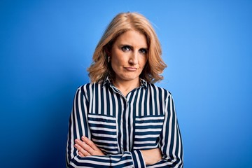 Middle age beautiful blonde woman wearing casual striped shirt standing over blue background...