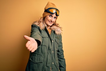 Middle age beautiful blonde skier woman wearing snow sportwear and ski goggles smiling cheerful offering palm hand giving assistance and acceptance.