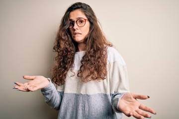 Fototapeta premium Young beautiful woman with curly hair wearing sweater and glasses over white background clueless and confused expression with arms and hands raised. Doubt concept.