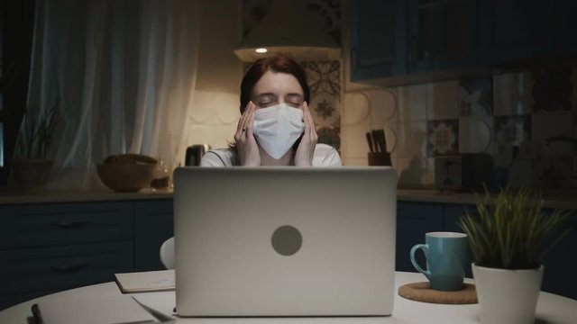 Young Girl In The Kitchen With Laptop. Young Woman Working Remotely At Home Because Of Pandemic. Woman In Medical Mask. Woman Tired. Her Head Hurts.