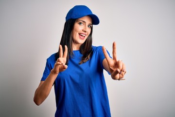 Young delivery woman with blue eyes wearing cap standing over blue background smiling looking to the camera showing fingers doing victory sign. Number two.