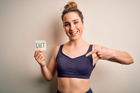 Young beautiful blonde sportswoman wearing sportswear holding reminder with diet message with surprise face pointing finger to himself
