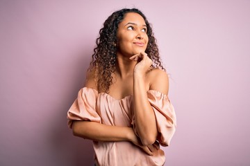 Young beautiful woman with curly hair wearing casual t-shirt standing over pink background with hand on chin thinking about question, pensive expression. Smiling and thoughtful face. Doubt concept.