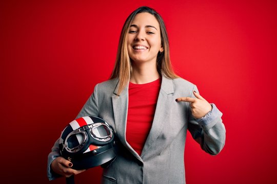 Young beautiful blonde motorcyclist woman holding motorcycle helmet over red background with surprise face pointing finger to himself