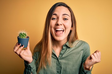 Young beautiful blonde woman with blue eyes holding small cactus pot over yellow bckground screaming proud and celebrating victory and success very excited, cheering emotion