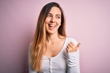 Young beautiful blonde woman with blue eyes wearing white t-shirt over pink background smiling with happy face looking and pointing to the side with thumb up.