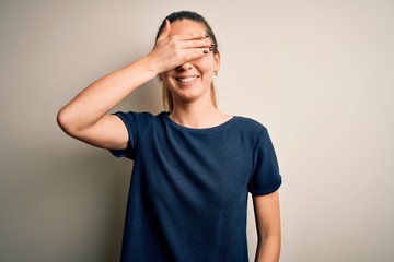 Young beautiful blonde woman with blue eyes wearing casual t-shirt over white background smiling and laughing with hand on face covering eyes for surprise. Blind concept.