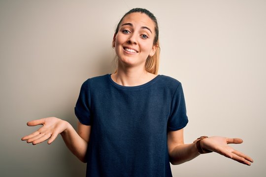 Young beautiful blonde woman with blue eyes wearing casual t-shirt over white background clueless and confused expression with arms and hands raised. Doubt concept.