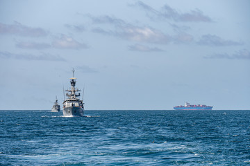 Two corvette warships sail together in the sea with container ship on the horizon