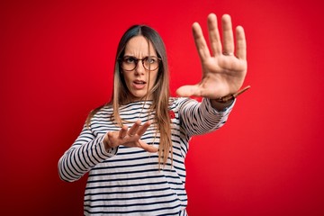 Young beautiful blonde woman with blue eyes wearing glasses standing over red background afraid and terrified with fear expression stop gesture with hands, shouting in shock. Panic concept.