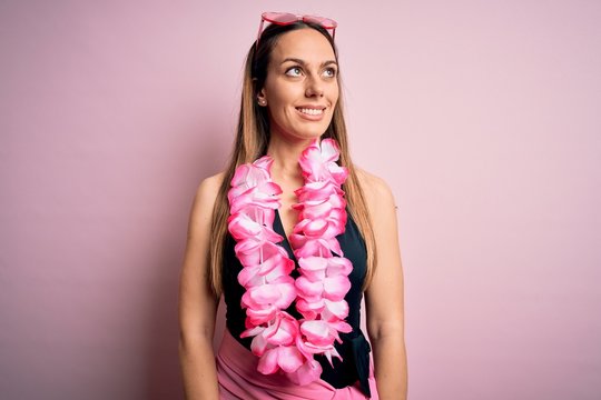 Young beautiful blonde woman wearing swimsuit and floral Hawaiian lei over pink background looking away to side with smile on face, natural expression. Laughing confident.