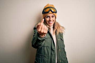 Young brunette skier woman wearing snow clothes and ski goggles over white background Beckoning come here gesture with hand inviting welcoming happy and smiling